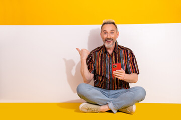 Excited Mature Man Holding Smartphone Pointing to the Side Against a Yellow Background Sitting...