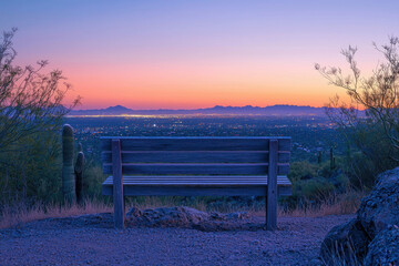 A wooden bench overlooks a stunning sunset over a city nestled in mountains.