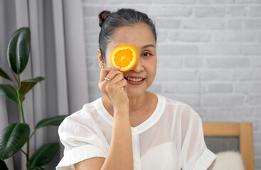 Senior asian woman enjoy a self-care moment with orange slice on her eye in a bright living room. Green plant accentuate the serene atmosphere.