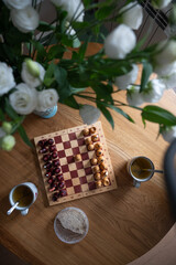 Chessboard with wooden pieces set up for a game, two teacups, a small bowl of sugar, and a vase of white flowers resting on a wooden table