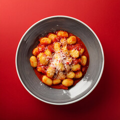 Delicious Italian gnocchi served with rich tomato sauce, grated cheese, and fresh parsley in a grey bowl on a vibrant red background.