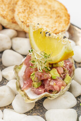 Close-up of a gourmet appetizer featuring tuna tartare with avocado and sprouts served in an oyster shell, topped with a sesame-crusted lemon wedge