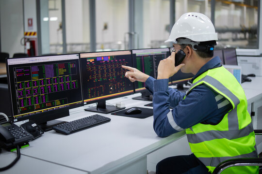 Technician in safety helmet uses computer systems to control and monitor substation operations with multiple screens displaying data and control panels in industrial setting