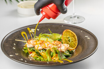 A close-up of a shrimp and vegetable salad being drizzled with bright yellow dressing, garnished with citrus and fresh greens