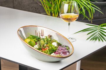 A fresh Greek-style salad with feta, red onion, and greens served in a ceramic bowl next to a glass of white wine on a modern white table
