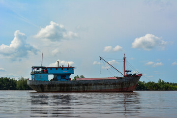 A large, weathered cargo ship with a light blue upper deck and a dark hull, navigating a wide, calm...
