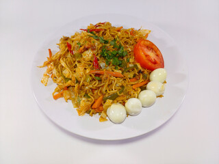 A vibrant plate of fried noodles (mie goreng) garnished with fresh vegetables, a tomato wedge, and several quail eggs shown on a white plate against a white set, defining a popular Asian culinary dish