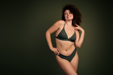 Confident young woman with curly hair posing in dark khaki lingerie against a green background