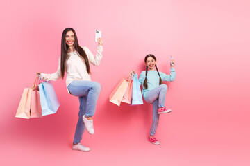Happy mother and daughter enjoying a shopping day with colorful bags and smartphones against a pink...