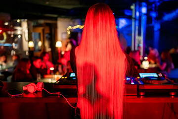 Female DJ with long hair mixing music in a dark nightclub, vibrant lights and energetic crowd in...
