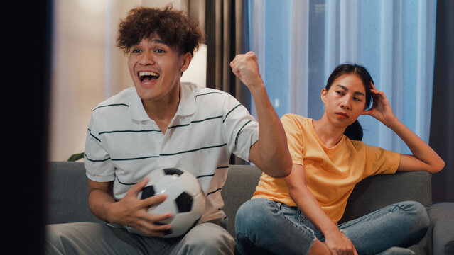 Asian new marriage couple sit on sofa husband excited cheering watching football sport game on television in living room at home. Young woman getting bored her partner watch online soccer on TV.