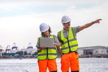 Two engineers wearing safety vests and helmets inspect port operations near the waterfront. They...