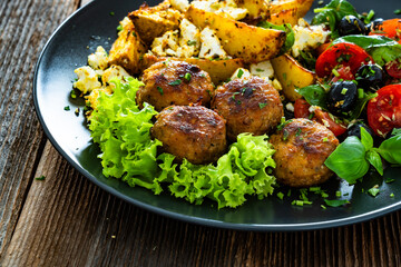 Greek style fried meatballs, fried potato wedges with feta cheese and tomato salad on wooden table	