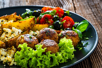 Greek style fried meatballs, fried potato wedges with feta cheese and tomato salad on wooden table	