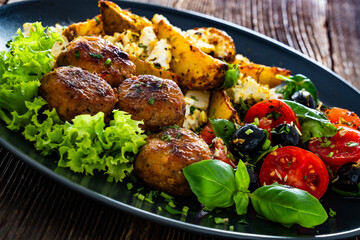 Greek style fried meatballs, fried potato wedges with feta cheese and tomato salad on wooden table	