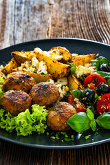 Greek style fried meatballs, fried potato wedges with feta cheese and tomato salad on wooden table	