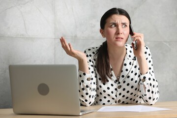 Young businesswoman with papers and laptop complaining to someone via phone at wooden table indoors