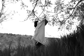 Black and white photograph of a girl holding an abstract-patterned carpet in wide open fields,...