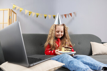 Birthday celebration. Little girl with piece of cake having video call via laptop on sofa at home
