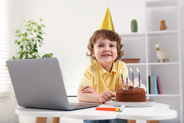 Birthday celebration. Little boy with cake having video call via laptop at table indoors