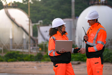 Two engineers in high-visibility safety gear and hard hats discuss project plans on a laptop in front of large spherical gas storage tanks at an industrial coastal facility.
