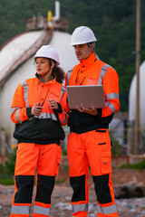Two engineers in high-visibility safety gear and hard hats discuss project plans on a laptop in front of large spherical gas storage tanks at an industrial coastal facility.
