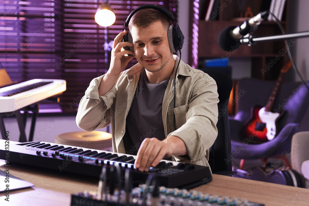 Poster smiling man in headphones working with midi keyboard at table in home studio