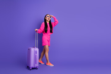 Young woman in stylish pink attire with luggage against a purple backdrop, showcasing trendy...