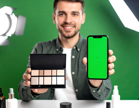 Smiling man holding makeup palette and smartphone with a green screen, showcasing beauty products.