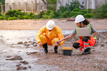 Two environmental engineers in safety gear work at a shoreline near gas storage tanks. One collects water samples, while the other records data on a laptop during site inspection.
