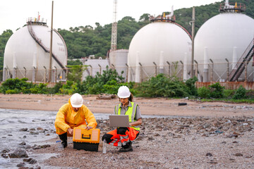 Two environmental engineers in safety gear work at a shoreline near gas storage tanks. One collects water samples, while the other records data on a laptop during site inspection.
