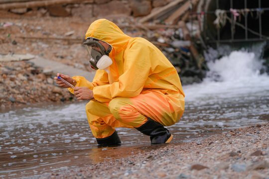Environmental field team in protective gear analyzes water samples on a polluted beach. One holds a laptop while the other handles vials for chemical testing of coastal pollution.