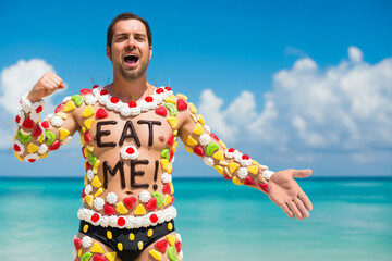 Fun, humorous photo of a funny young man on the beach whose body is decorated with candied fruit and whipped cream, looking ridiculous with "EAT ME" written in chocolate sauce on his torso