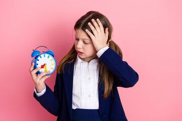 Young schoolgirl holding a blue alarm clock on a pink background, showing concern and urgency, dressed in formal school uniform