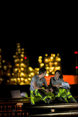 Two industrial workers take a break during the night shift at a construction site, sitting on steel...