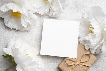 Blank wedding invitation mockup with copy space surrounded by white peonies and a wrapped gift,...