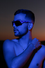 Stylish man illuminated by vibrant red lighting at sunset wearing sunglasses and silver chains
