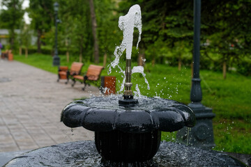 A close-up view of a water jet emerging from a metal fountain in a summer city park, with benches and a paved alley in soft focus, evoking cool relaxation. Photo. High resolution