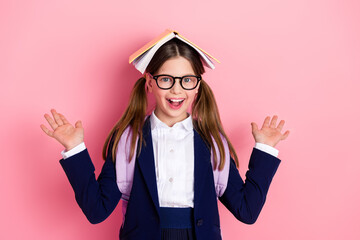 Cheerful schoolgirl with a book on her head poses enthusiastically on pink background, showcasing...