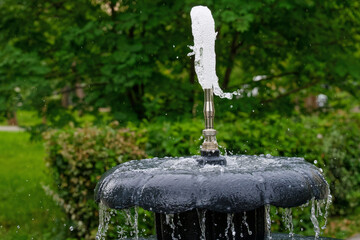 A dynamic water jet spurting from a metal fountain nozzle in a lush summer city park, capturing droplets in sharp focus and conveying a refreshing ambiance. Photo. High resolution