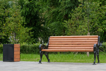 Empty wooden bench and litter bin on paved walkway in green summer park. Ideal resting place in urban environment. Photo. High resolution