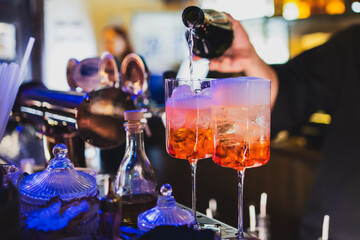 Bartender pouring cocktails into two glasses with ice at a vibrant bar counter, surrounded by colorful lights, bottles, and bar tools, capturing nightlife and mixology in action.