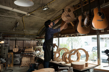 Carpenter hanging unfinished wooden guitar on rack in workshop filled with tools, frames, and musical instruments