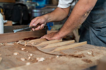 Close-up of craftsman's hands chiseling wood inside guitar body with precision on dusty workbench in workshop