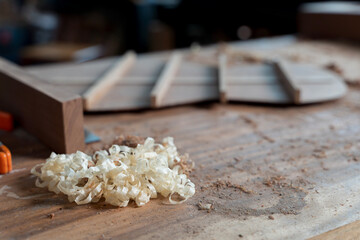 Close-up of wood shavings on dusty workbench in carpentry studio with blurred unfinished guitar part in background