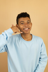 Vertical image of a cheerful brown-skinned Latino teenager wearing a pastel blue sweatshirt making a call gesture with his hand on a light brown background with copy space
