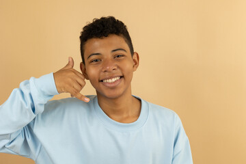 Cheerful brown skinned Latino teenager wearing a pastel blue sweatshirt making a call gesture with his hand on a light brown background with copy space