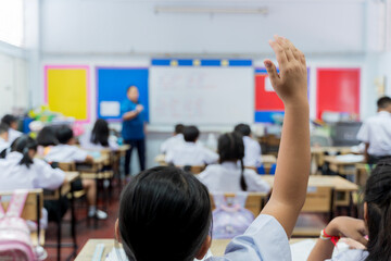 Student raising hand to answer question in active classroom with teacher and children in school uniform learning