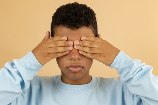 Dark skinned Latino teenager wearing a pastel blue sweatshirt, covering his eyes with his hands on a light brown background, concept of hiding sight