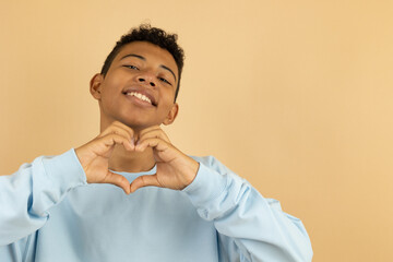 Cheerful brown skinned teenager wearing a pastel blue sweatshirt making a heart gesture with her hand on a light brown background with copy space, young man expressing feeling of love with his hands